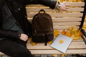 person sat on bench with bag and laptop and autumnal leaves 